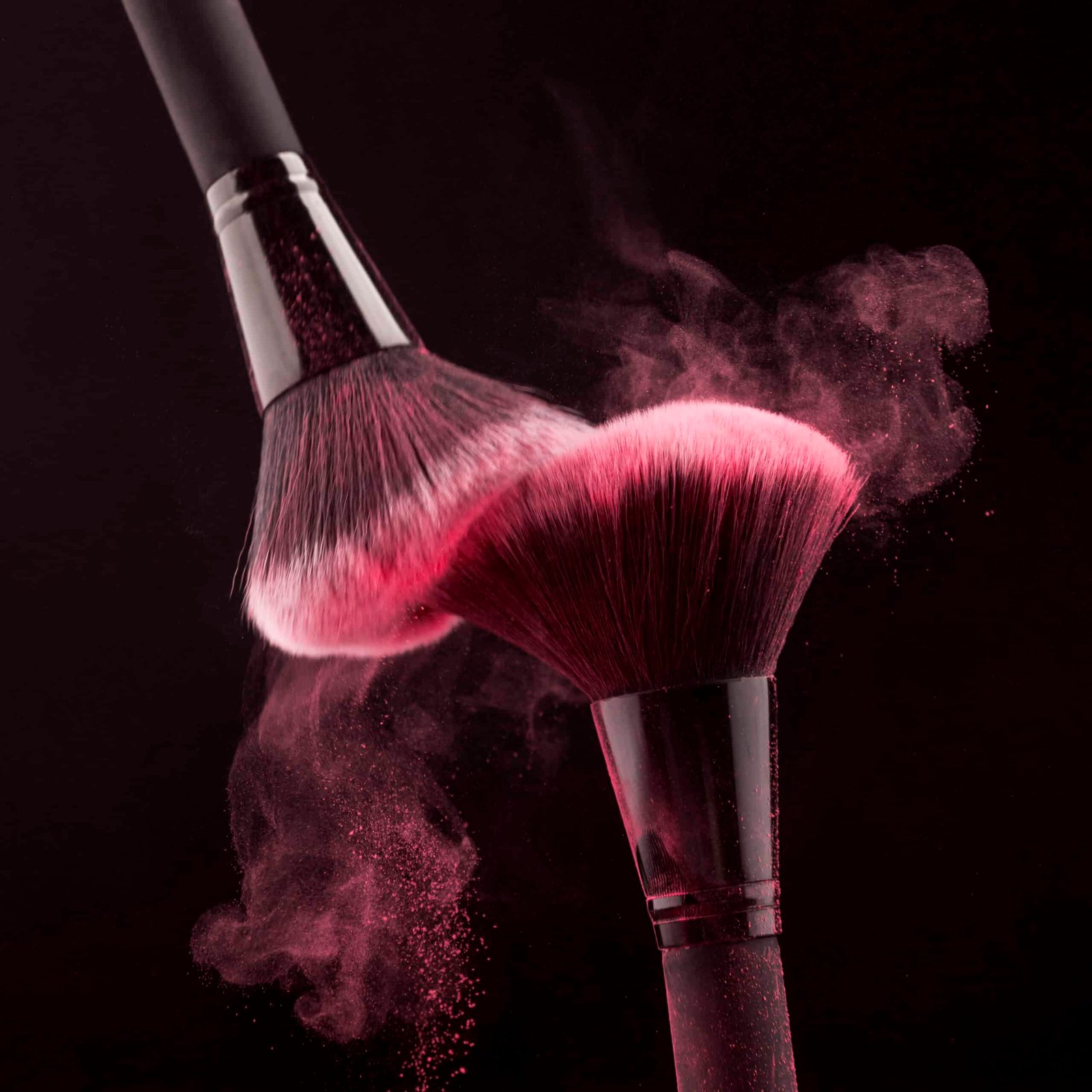 Deux pinceaux s'effleurant libèrent une poudre de couleur rose dans une lumière tamisée, capturant la beauté de l'art du maquillage.